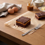 Two billionaire brownies on parchment paper on a wooden table, showing the brownie base, caramel layer and chocolate top, with a fork in the foreground and eggs, a jar of caramel and butter in the background.