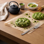 Two green matcha cookies topped with white chocolate pieces on parchment paper, set on a wooden table with a black teapot, a cup of green tea, a bowl of matcha powder, a bamboo whisk, and a silver fork.