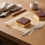 Two Millionaire shortbreads on parchment paper on a wooden table, with a fork, cloth, brush, and small jar in the background.