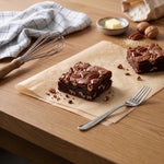 Two Nutella and Pecan brownies on parchment paper on a wooden table, styled with a fork, whisk, butter, and walnuts.